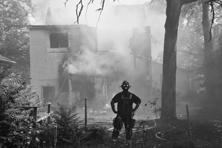 A firefighter stands in front of a smoking, partially burnt house amidst greenery. Day outdoors.